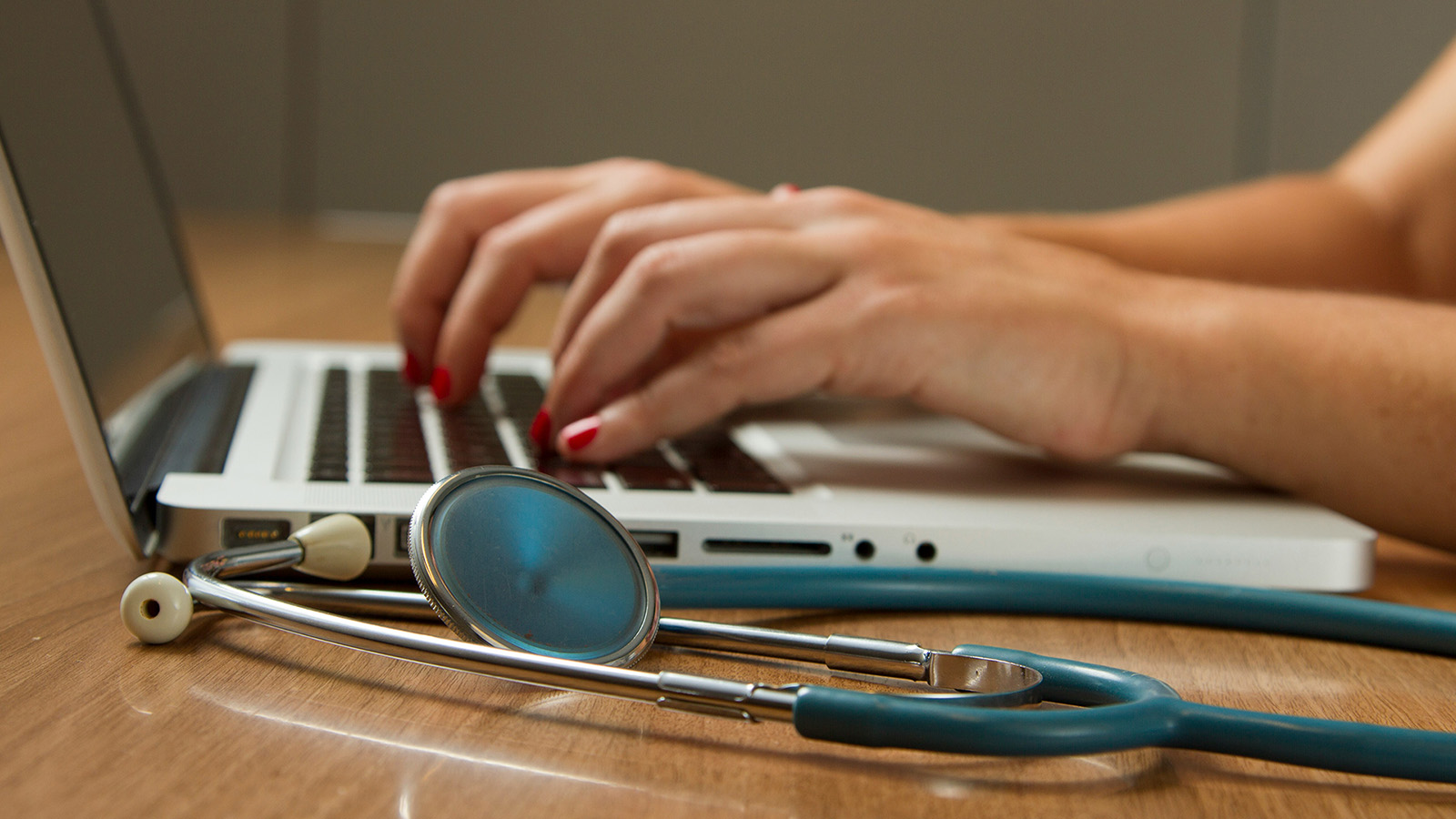 Person typing on a laptop with a stethoscope lying on the wooden desk