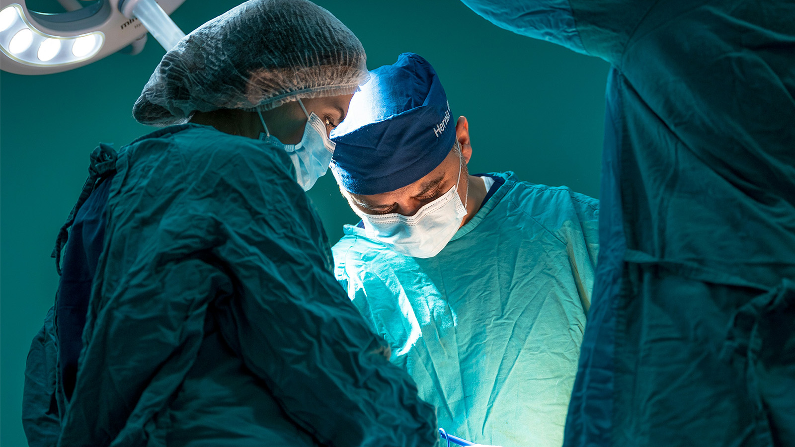 Surgeons wearing masks, gowns, and caps perform a medical procedure with precision under bright surgical lights in an operating theatre.