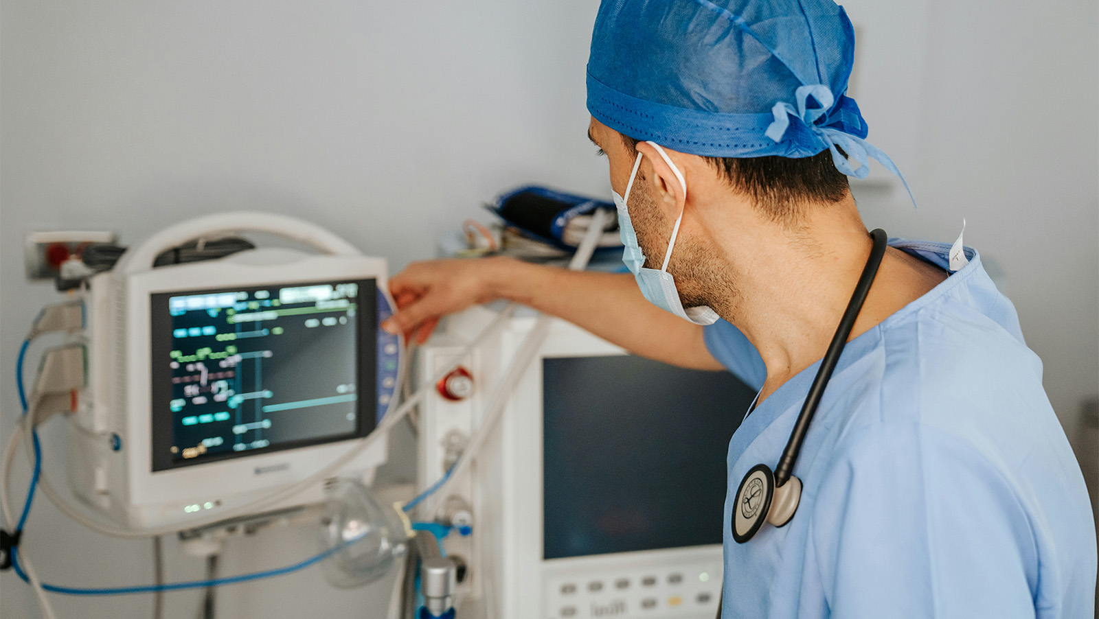 A medical professional in scrubs and mask monitors patient vital signs on a screen