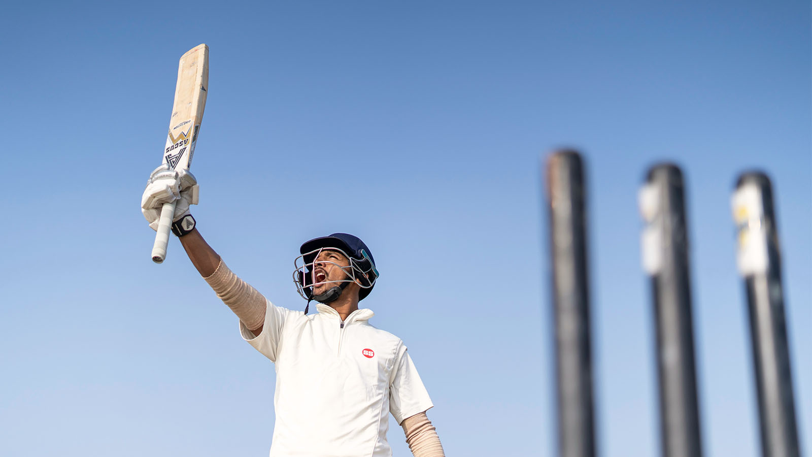 A cricket player wearing a helmet and white kit raises his bat and shouts in celebration