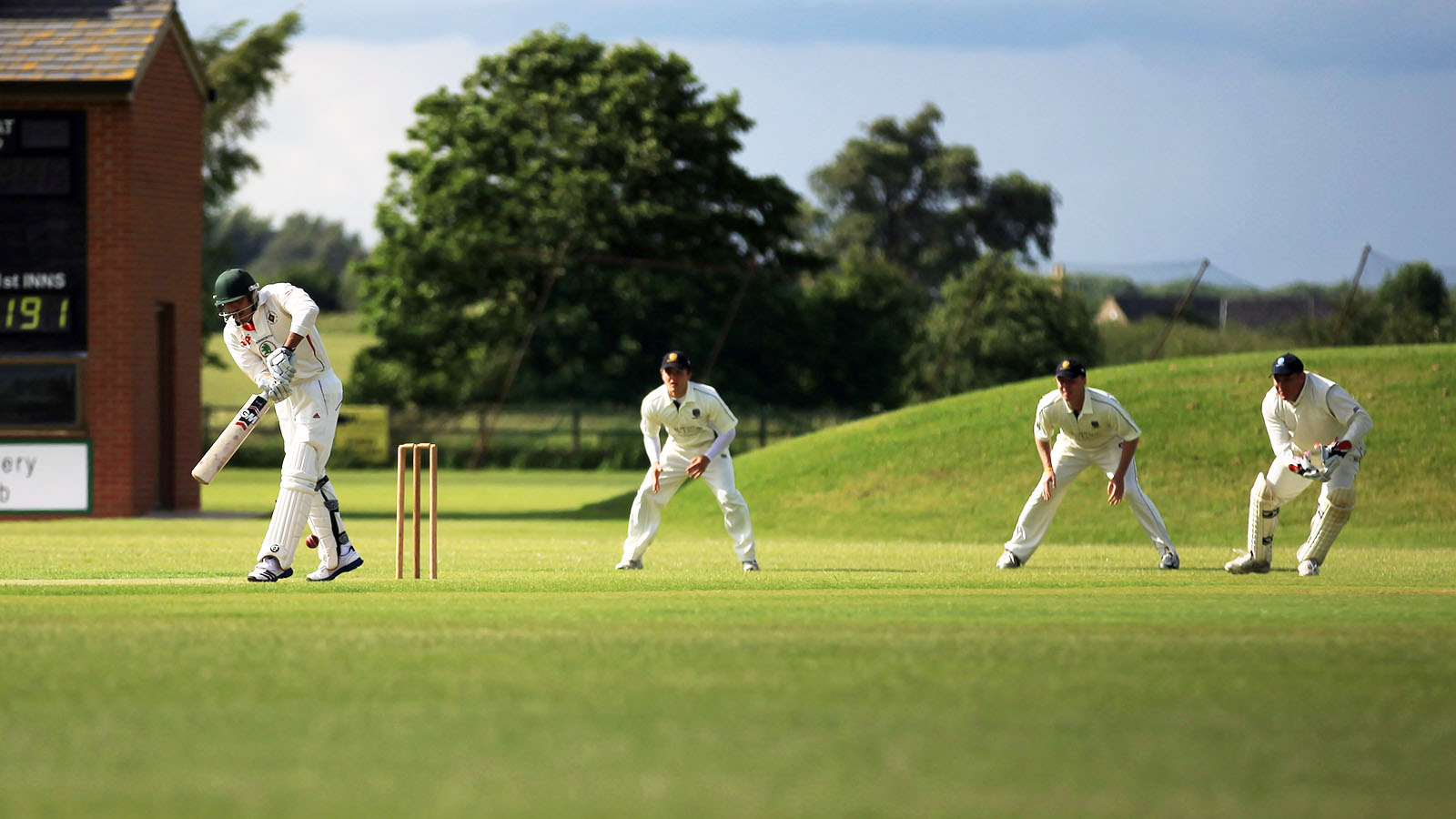 A cricket batsman defends a ball while three fielders and a wicket-keeper stand behind the stumps