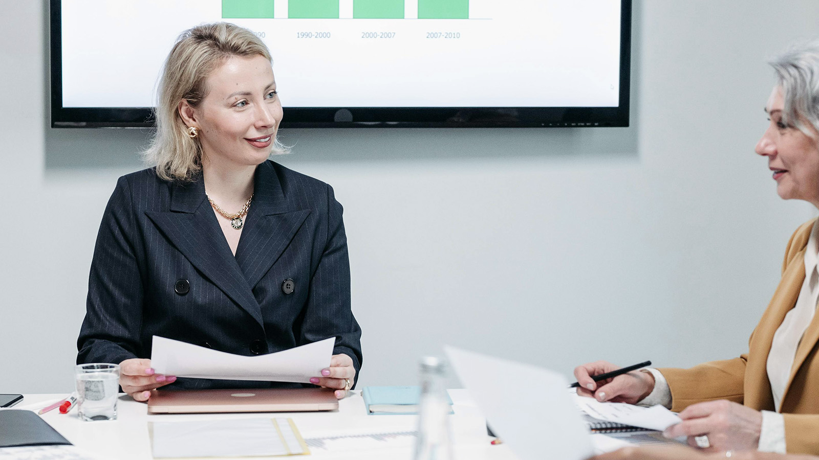 Two women in business attire sit at a desk with documents