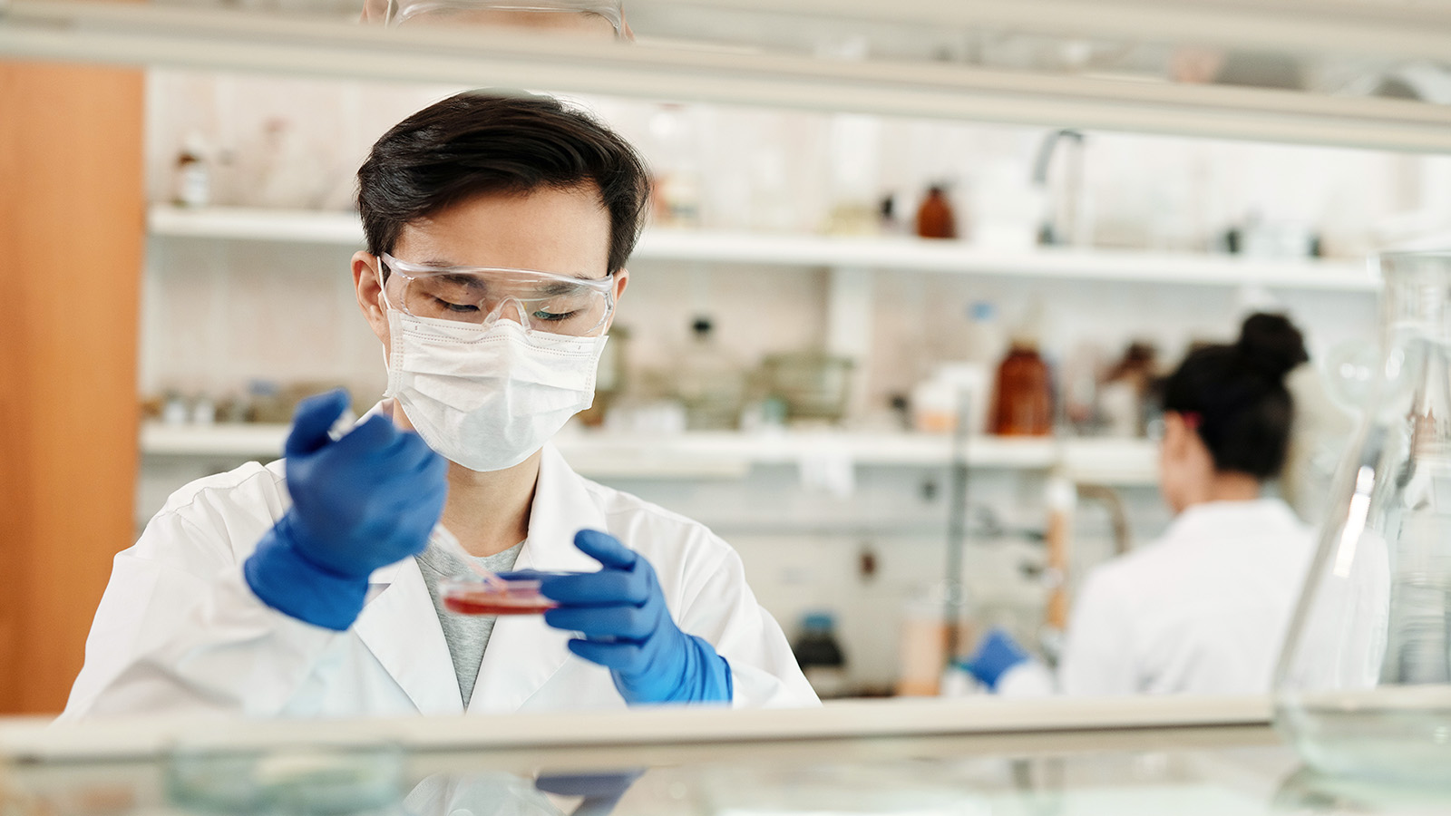 A scientist wearing a mask, goggles, and gloves uses a pipette with a petri dish in a laboratory
