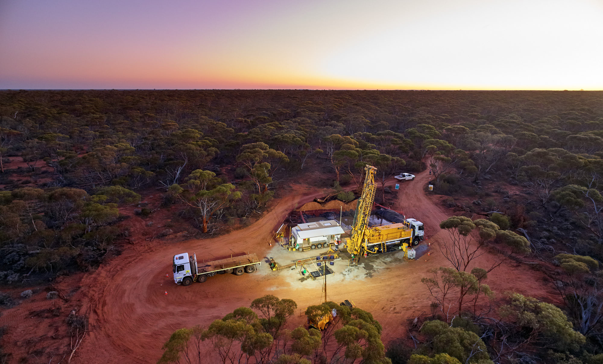 Aerial view of a drilling rig and lorries operating on a cleared dirt site surrounded by dense bushland at sunset, capturing the dynamic scene of energy and mining activity.