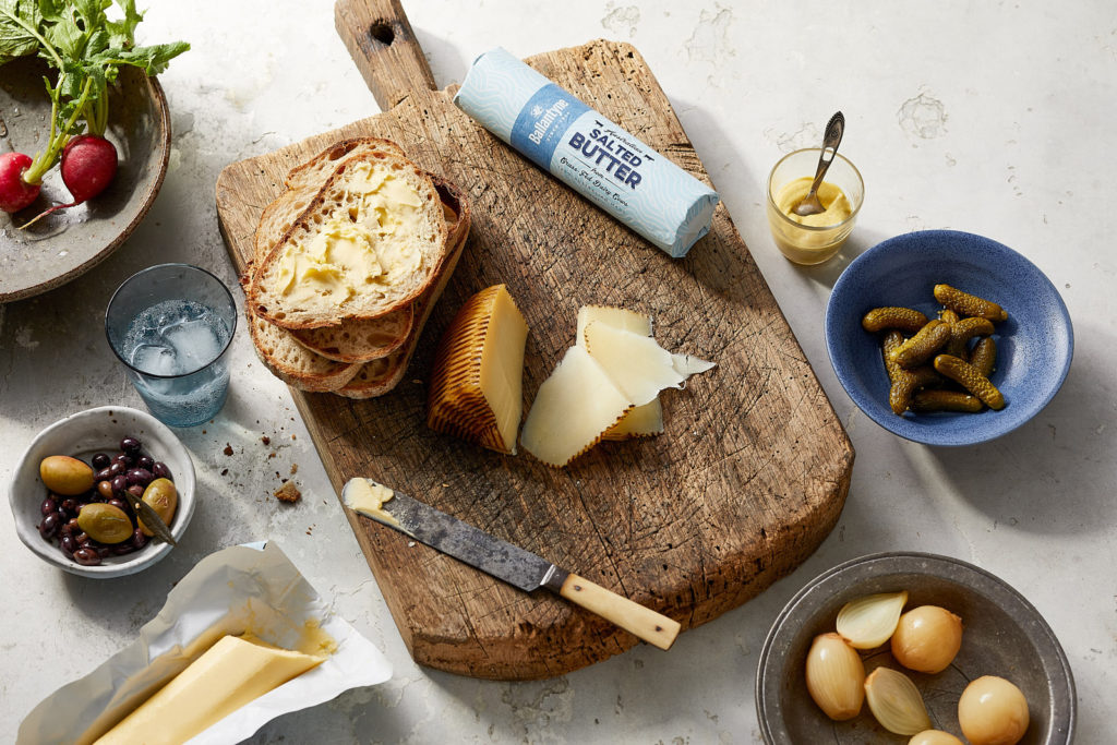 A wooden board with buttered bread, sliced cheese, and a knife, surrounded by bowls of pickles, olives, onions, mustard, and a pack of salted butter showcases the delicious results of quality food manufacturing.