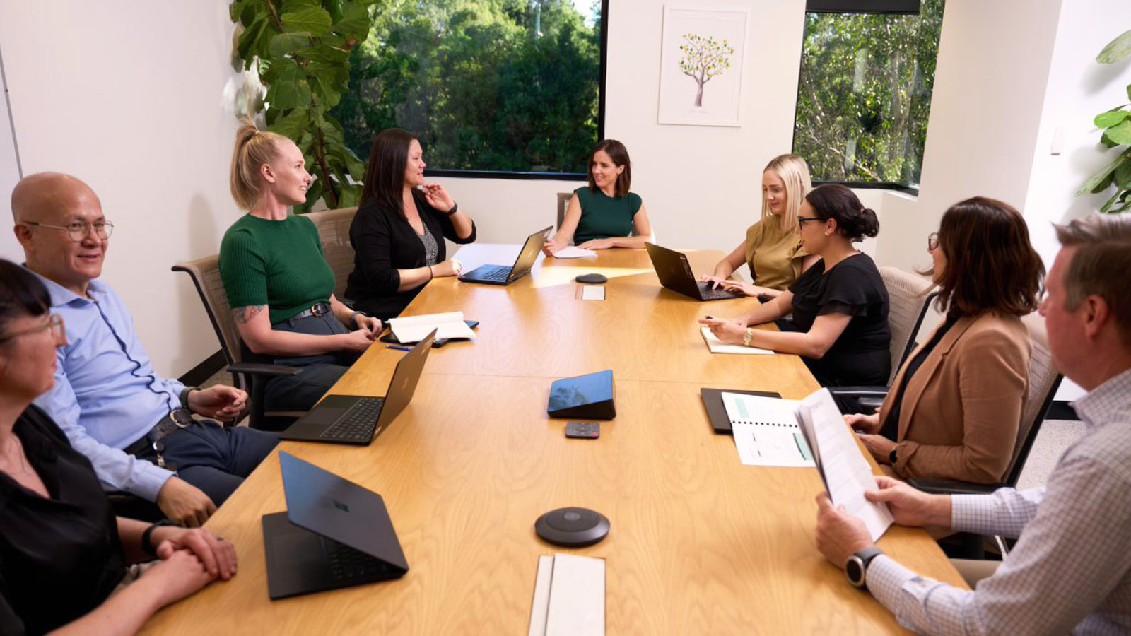 Ten people sit around a conference table with laptops and papers, engaged in a meeting at Integria Healthcare, using Workday Adaptive Planning for better planning in a bright office with large windows and plants.