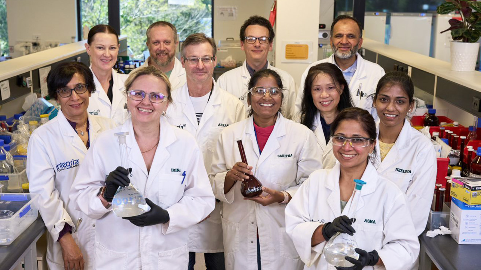 A group of scientists in lab coats stand together in a laboratory, some holding glassware, with shelves of equipment and supplies in the background—demonstrating Integria Healthcare’s commitment to better planning and innovation.