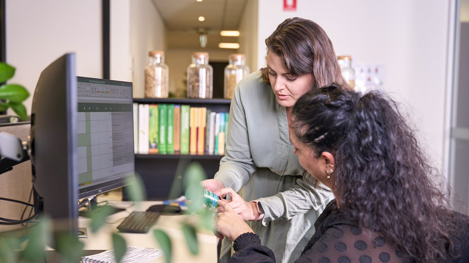 Two women collaborate at a desk; one is seated using a computer with Workday Adaptive Planning on screen, whilst the other stands beside her, pointing at something on a mobile for better planning.
