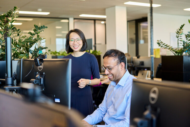 Two colleagues, including Nuwan Fernando, work together at computer desks in a modern office with large windows and plants in the background.