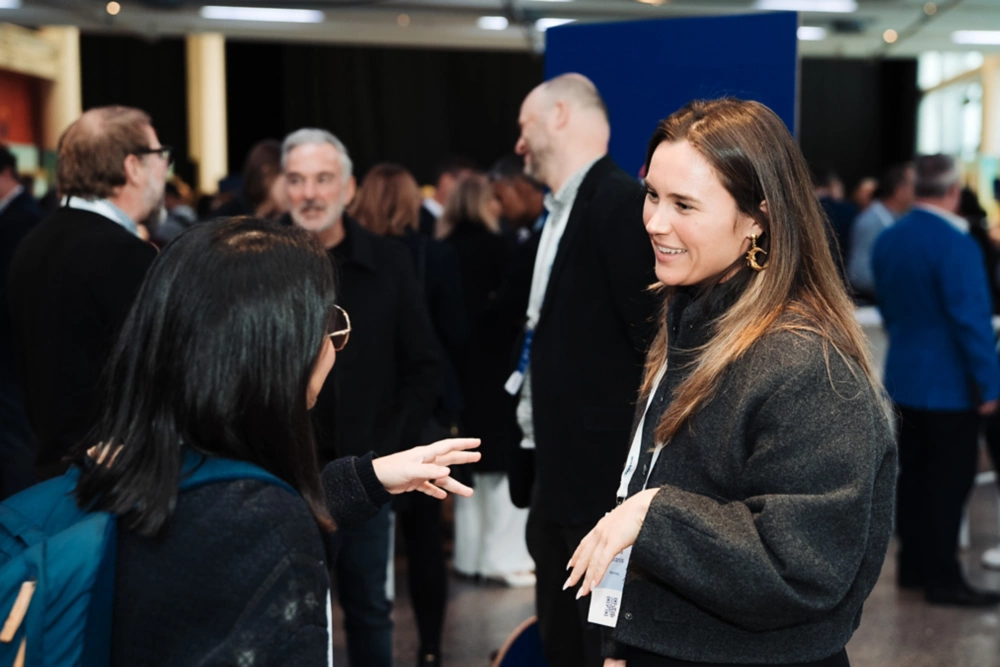 Two women converse and smile at a crowded indoor event, surrounded by others engaged in lively discussions about AI and ML innovations.