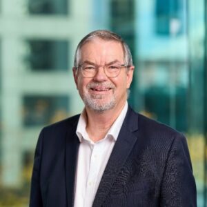 A middle-aged man with glasses, grey hair, and a beard, wearing a dark suit and white shirt, smiles confidently in front of a blurred office window—reflecting expertise in Aged Care Finance Sydney.