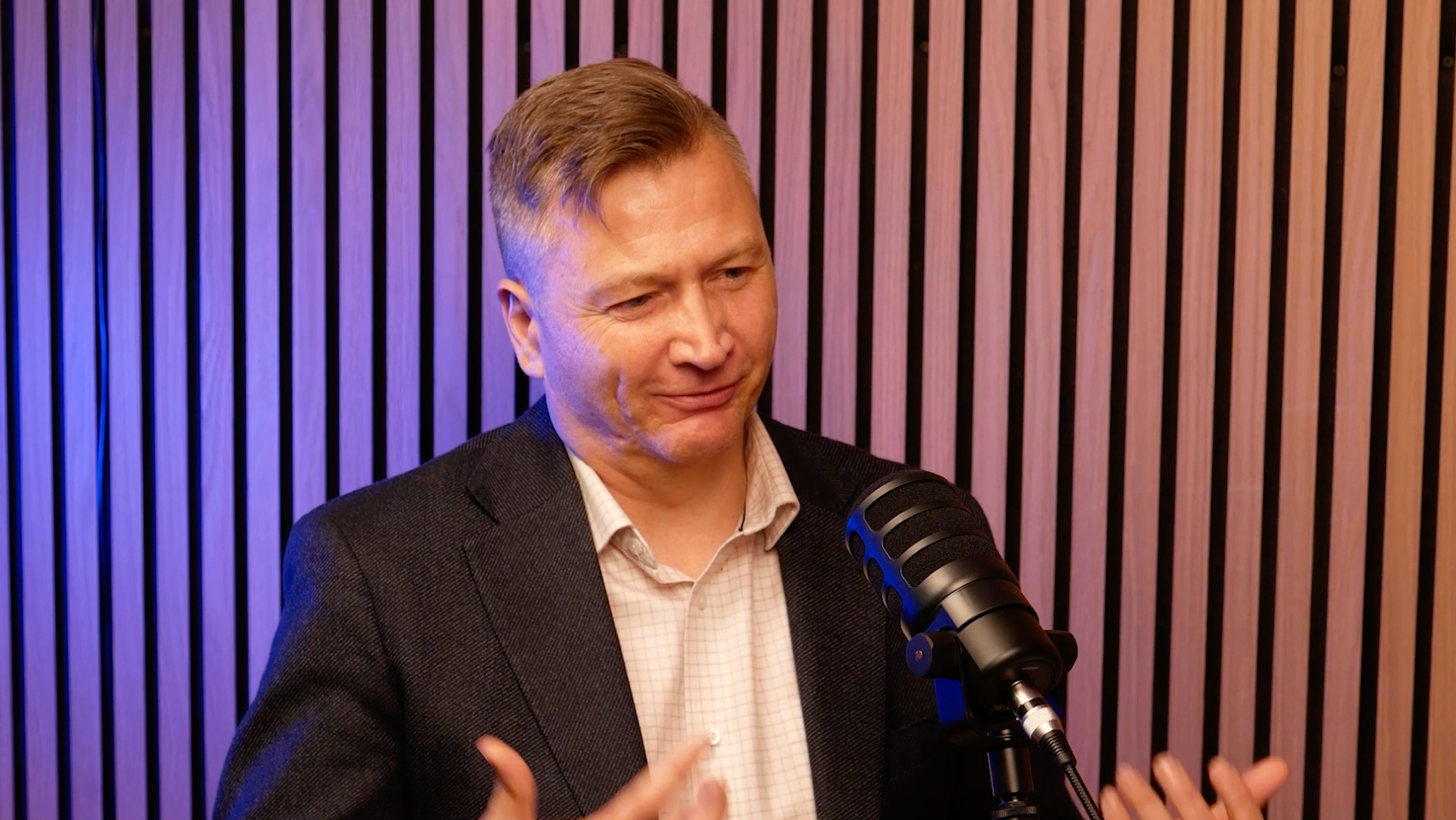 A man in a blazer speaks into a microphone in front of a striped wooden background, gesturing with his hands as he discusses the benefits of partnering with QMetrix for seamless Workday Adaptive Planning implementation.