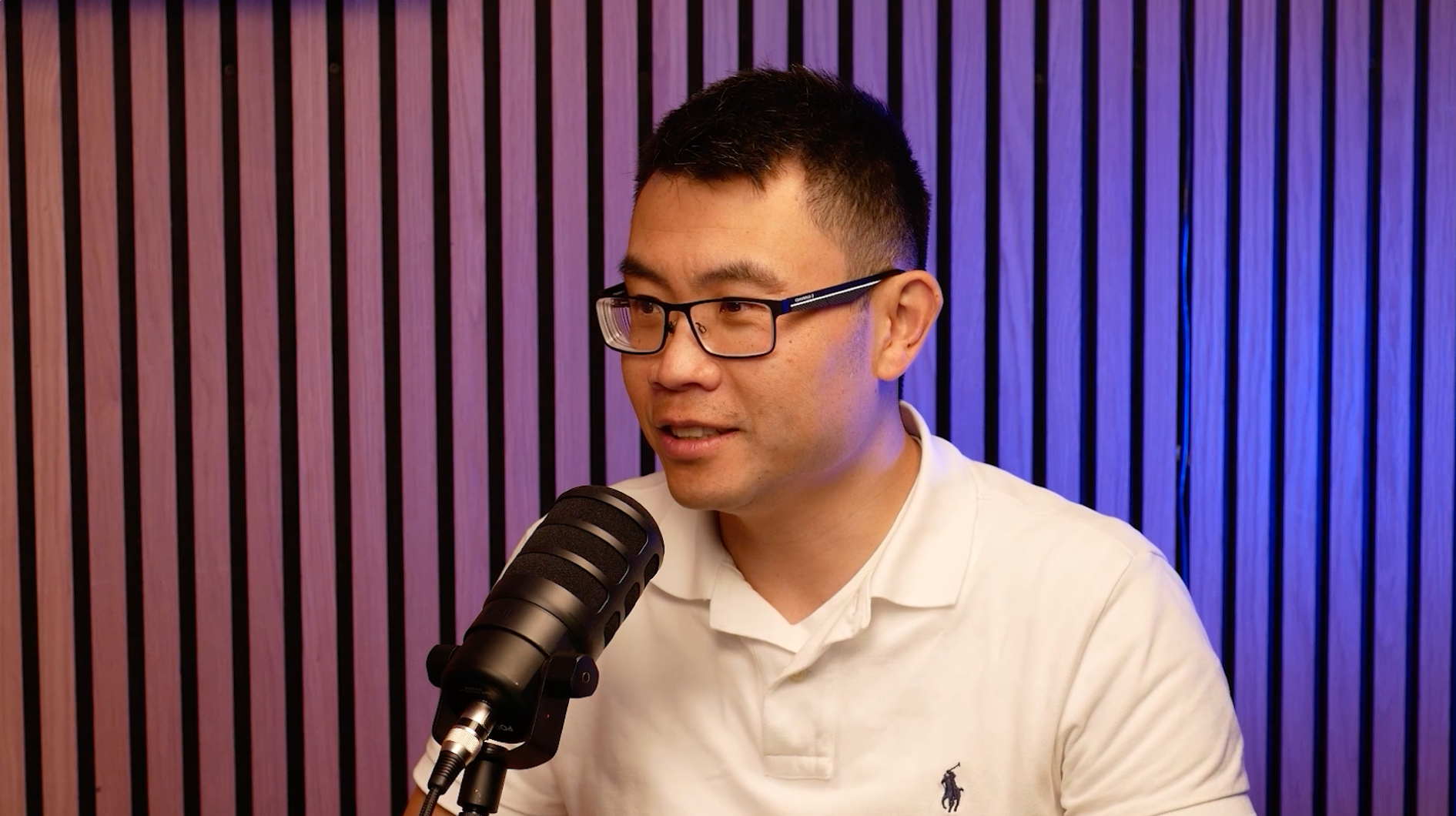 A man in glasses and a white polo shirt speaks into a black microphone, seated in front of vertical wooden slats with purple lighting, discussing how QMetrix serves as an implementation partner for Workday Adaptive Planning.