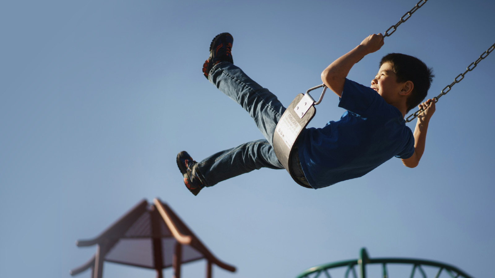 A boy in a blue shirt swings high on a playground swing set, with his legs outstretched and playground structures visible in the background under a clear sky—capturing the effortless momentum of a fast FP&A implementation.