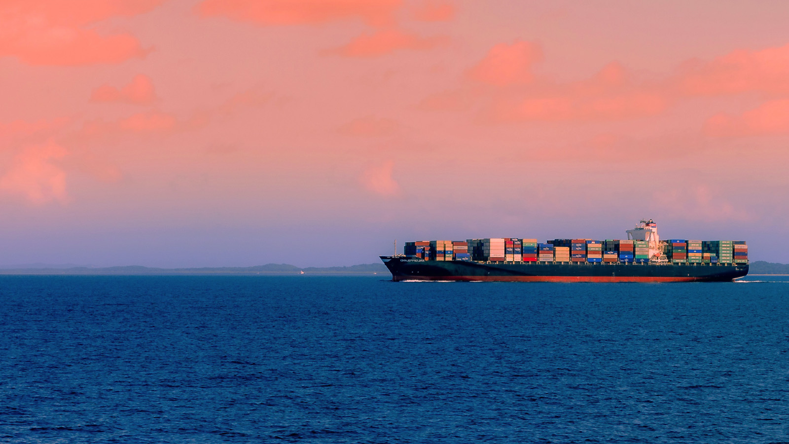 A large cargo ship loaded with colourful shipping containers sails across a calm blue sea under a pink sky